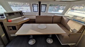 Et bådeinteriør med et centralt bord, polstrede siddepladser, et kontrolpanel og vinduer med udsigt over en lystbådehavn.