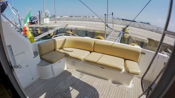 Interior de un barco con zona de asientos acolchada, cubierta de madera y vista al muelle y al agua.