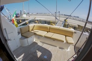 A boat's interior with a cushioned seating area, a wooden deck, and a view of the dock and water.