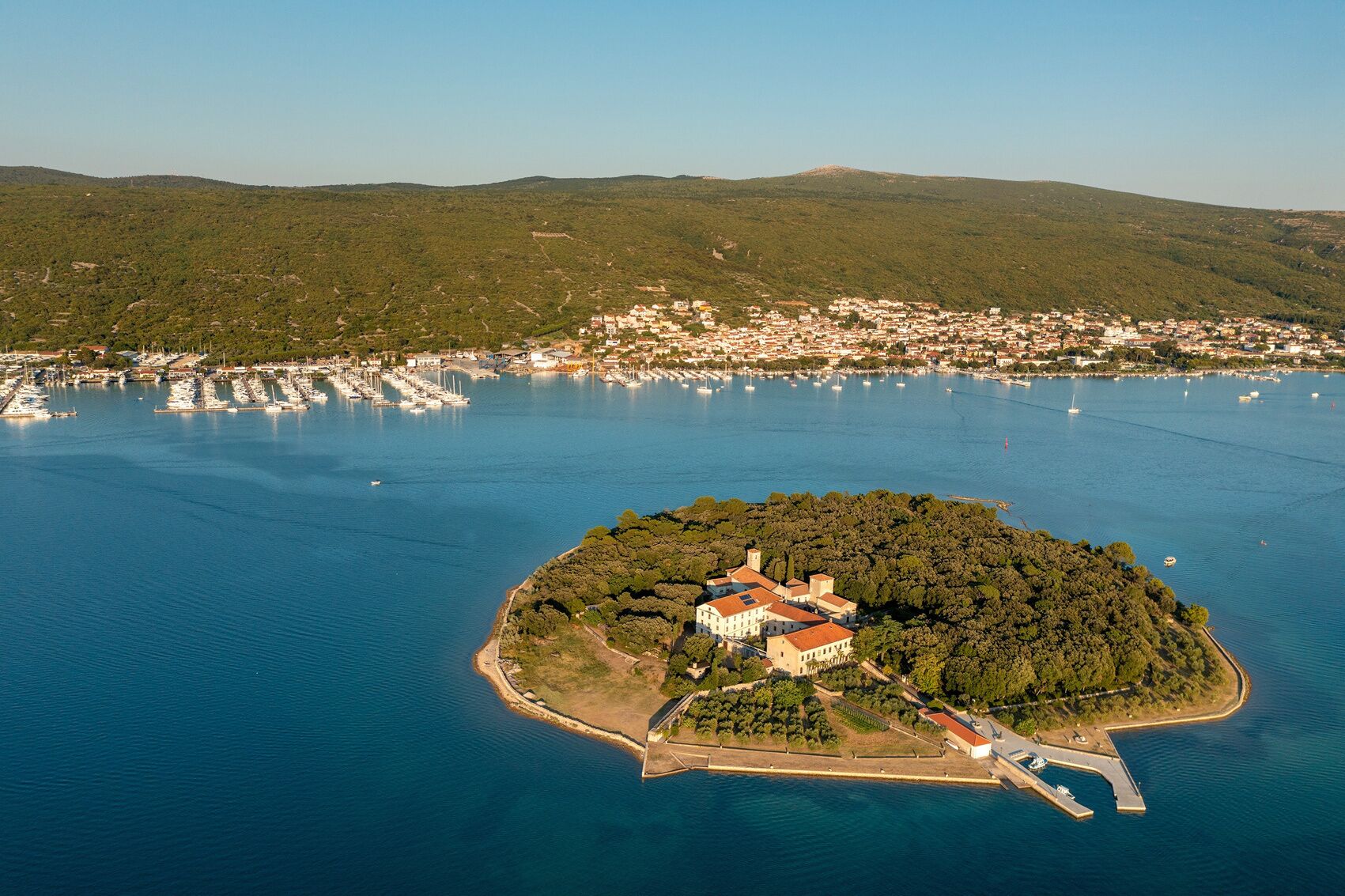 An aerial view of a coastal town with a prominent island featuring a large building with a red roof, surrounded by water and greenery.
