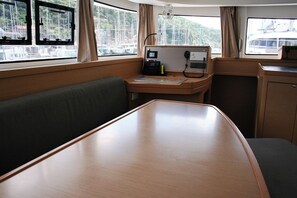A cabin with a wooden table, a cushioned bench, a small desk with a phone and a lamp, and windows with curtains. - Dubrovnik Delight: A Sun-Kissed Sailing Experience (Komolac)