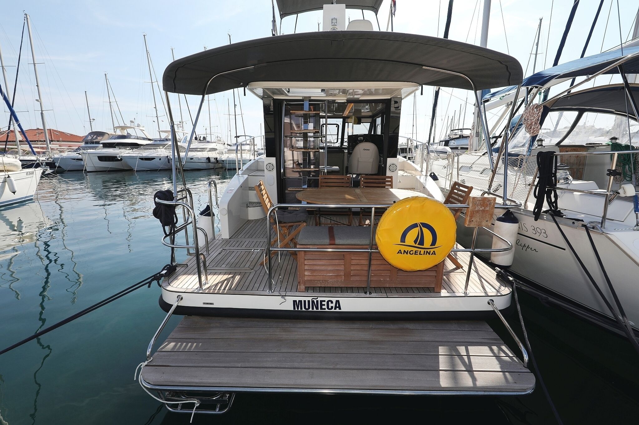 Ein Yachthafen mit einem Boot namens 'MUNÉCA', das einen überdachten Deckbereich mit Sitzgelegenheiten und einem Tisch bietet.