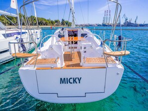 A sailboat named 'MICKY' with a wooden deck and metal railings, docked in a marina with clear blue water and industrial structures in the background.