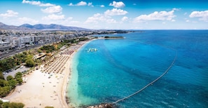Un paysage urbain côtier avec une plage, une eau d’un bleu limpide et une chaîne de montagnes à l’horizon.