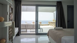 Ein Schlafzimmer mit einem großen Bett, einer Steinsäule, einem Balkon mit Meerblick und einer Sitzecke mit Tisch und Stühlen.