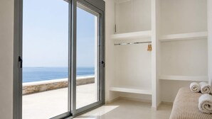Ein Zimmer mit einer großen Schiebeglasdoor zum Balkon mit Meerblick. Weiße Regale und ein Bett mit aufgerollten Handtüchern.