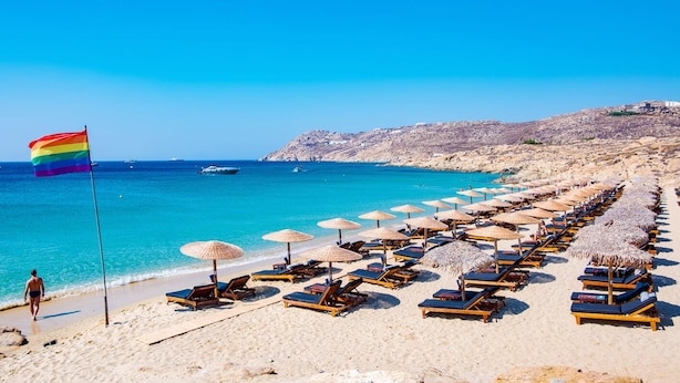 A beach with rows of sun loungers and umbrellas, a flag, and a clear blue sea. - Sun-Kissed Sailing Adventure in Islas Cícladas, Mykonos (Mykonos)