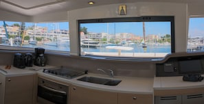 A compact kitchen with a stove, sink, and coffee maker, overlooking a marina with boats and buildings.