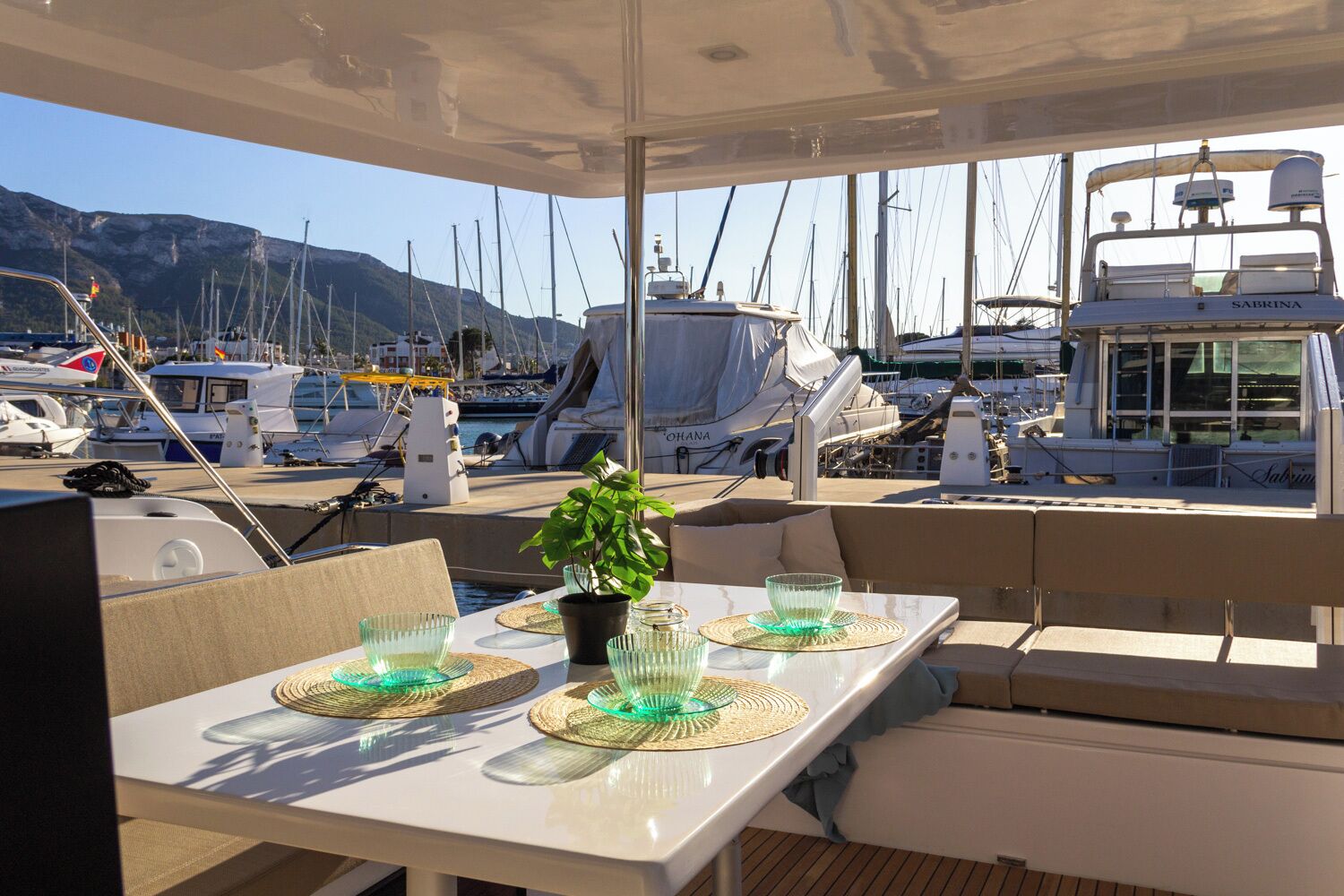 Convés de barco com mesa posta para refeição, rodeada por cadeiras e um vaso de planta.