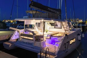 Eine Yacht namens 'Flor de Thalía' liegt in einer Marina vor Anker, mit 'AVENTURA'-Branding, einem überdachten Sitzbereich und einem kleinen Tisch mit einer Pflanze.