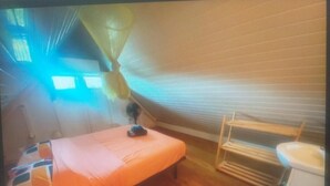 Ein kleines Zimmer mit einem Einzelbett, Holzboden, einem Nachttisch, einem Stuhl, einem Fenster mit Vorhängen und einem weißen Waschbecken.