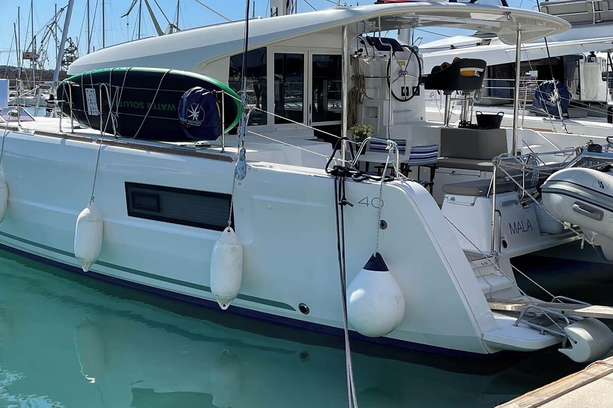 A white yacht docked at a marina with a covered deck area, a water-solvent buoy, and a winch labeled 'MALA'.