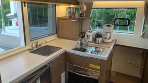 Cocina compacta con pileta, estufa y horno, una ventana con vista a follaje, y una encimera con diversos objetos.