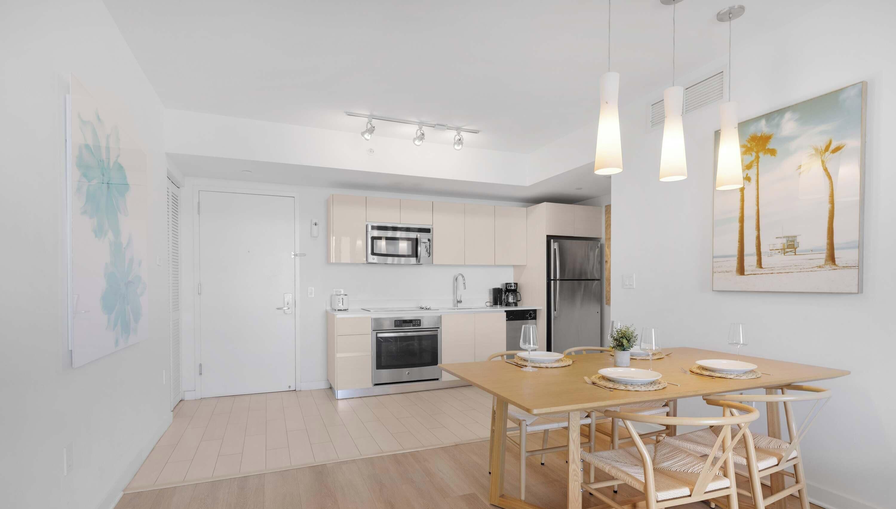 Una cocina moderna con electrodomésticos de acero inoxidable, un comedor con mesa y sillas de madera, y un cuadro con temática de playa en la pared.