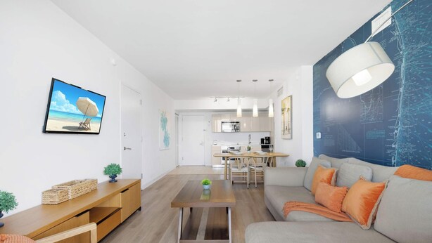 A modern living room with a sofa, coffee table, and a dining area with a kitchen in the background. - Spacious 1BR at Beachwalk (Hallandale Beach)