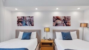 Ein Hotelzimmer mit zwei Betten, weißer Bettwäsche, blauen Kissen, einem Nachttisch mit Lampe und zwei an der Wand befestigten Kunstwerken.