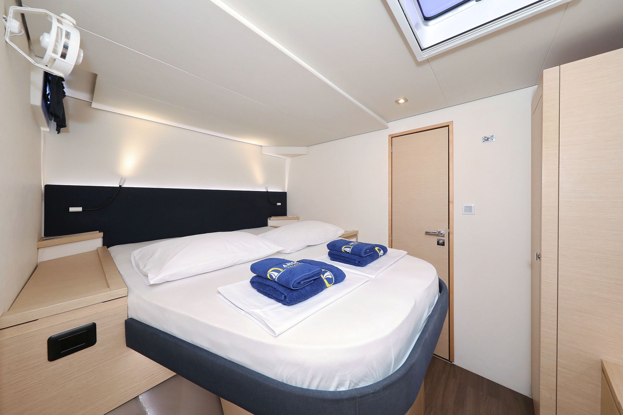 Cama bem arrumada com lençóis brancos e azuis, mesa de cabeceira com abajur e um guarda-roupa.