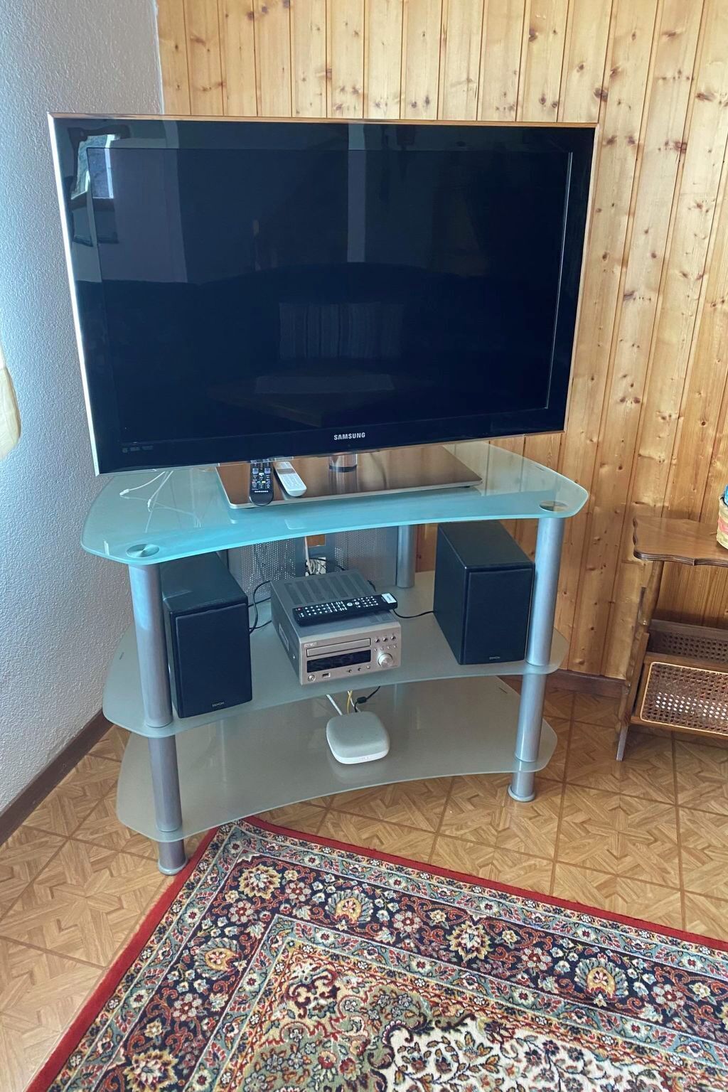 Un téléviseur à écran plat posé sur un meuble télé en verre, avec une télécommande, une chaîne stéréo et un tapis décoratif.
