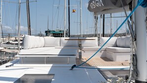 A yacht deck with white cushioned seating, a wooden deck, and multiple masts in the background. - Sun-Kissed Sailing: A Luxurious Beach Experience in Valencia (Denia)