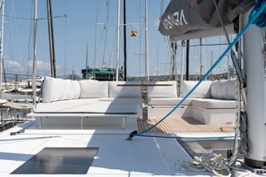 Ein Yachttdeck mit weißen, gepolsterten Sitzgelegenheiten, einem Holzboden und mehreren Masten im Hintergrund.