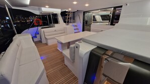 A yacht interior with a seating area, a table, and a view of the city at night. - Sun-Kissed Sailing: A Luxurious Beach Experience in Valencia (Denia)