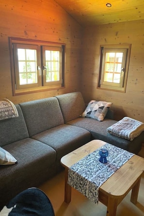 A cozy cabin interior with a sectional sofa, a wooden coffee table, and two windows with curtains.