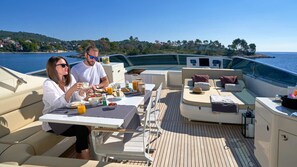 A boat with a dining area, lounge seating, and a view of the sea and hills.