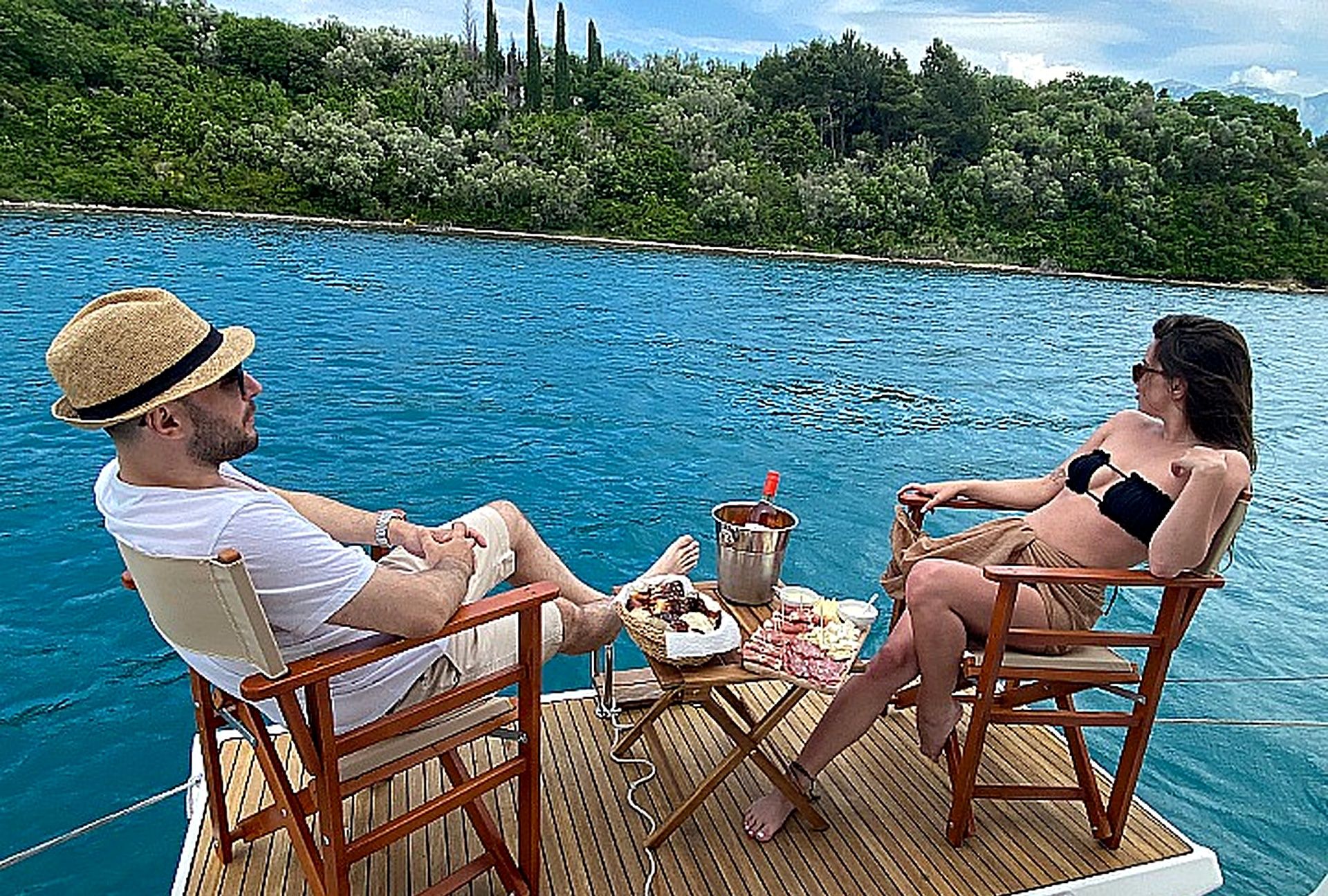 Dos personas sentadas en sillones de cubierta mirando una piscina, con una mesa entre ellas que sostiene comida y bebidas.