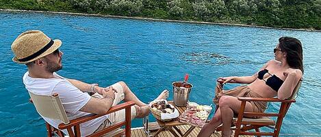 Two people seated on deck chairs facing a pool, with a table between them holding food and drinks.