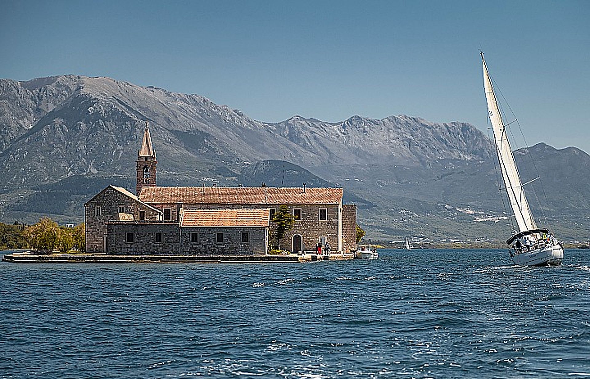 A stone building with a bell tower by a body of water, with a sailboat in the background.