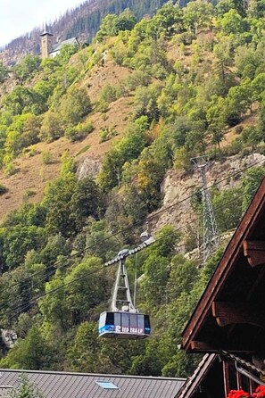 Eine Kabseilbahn mit einer Gondel mit der Aufschrift 'Bied Eralp', die über einer gebirgigen Landschaft mit dichter Vegetation und einer Kirche auf einem Hügel schwebt.
