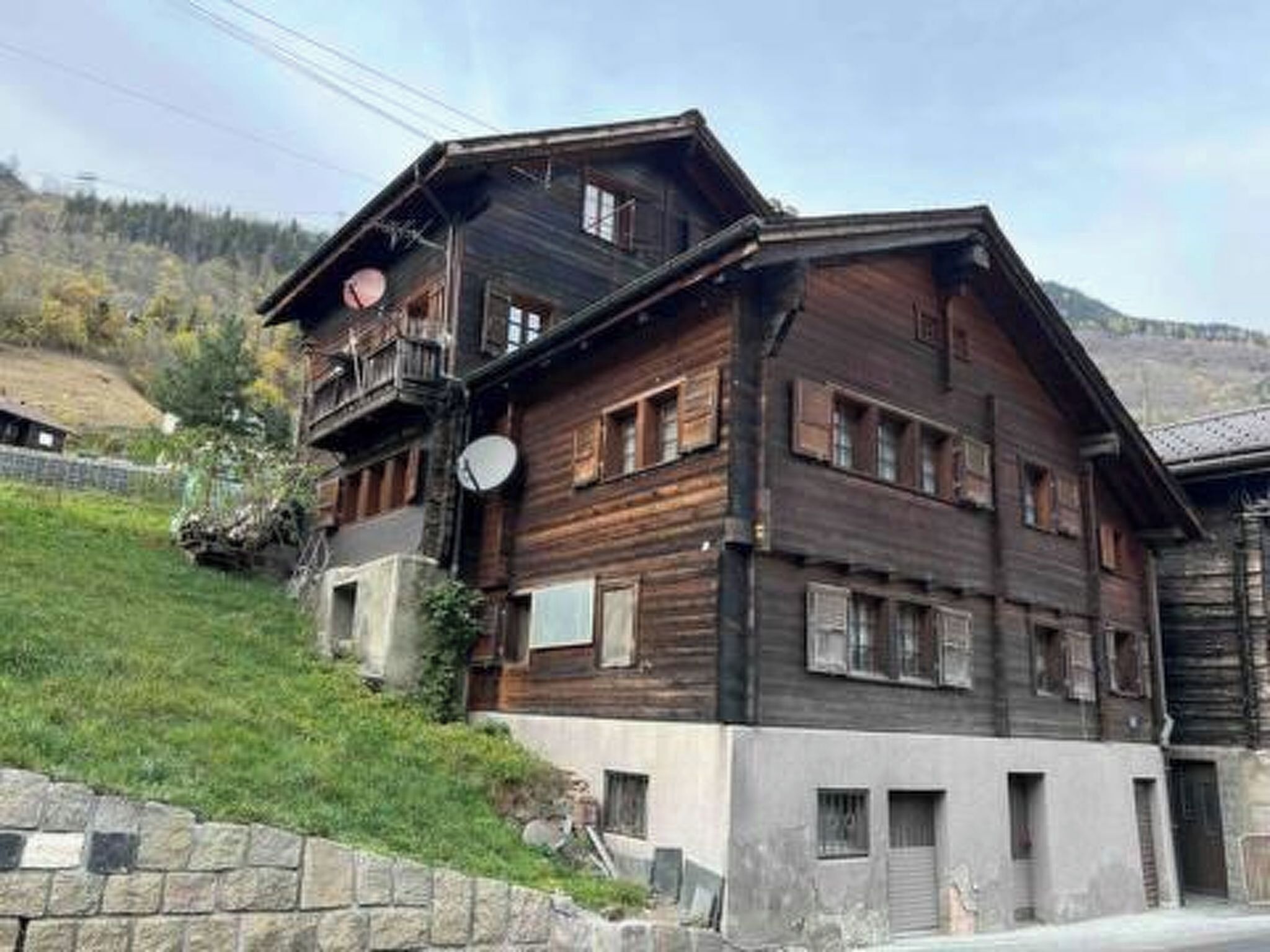 A multi-story wooden building with balconies, a stone wall, and a hilly background.