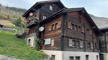 A multi-story wooden building with balconies, a stone wall, and a hilly background.