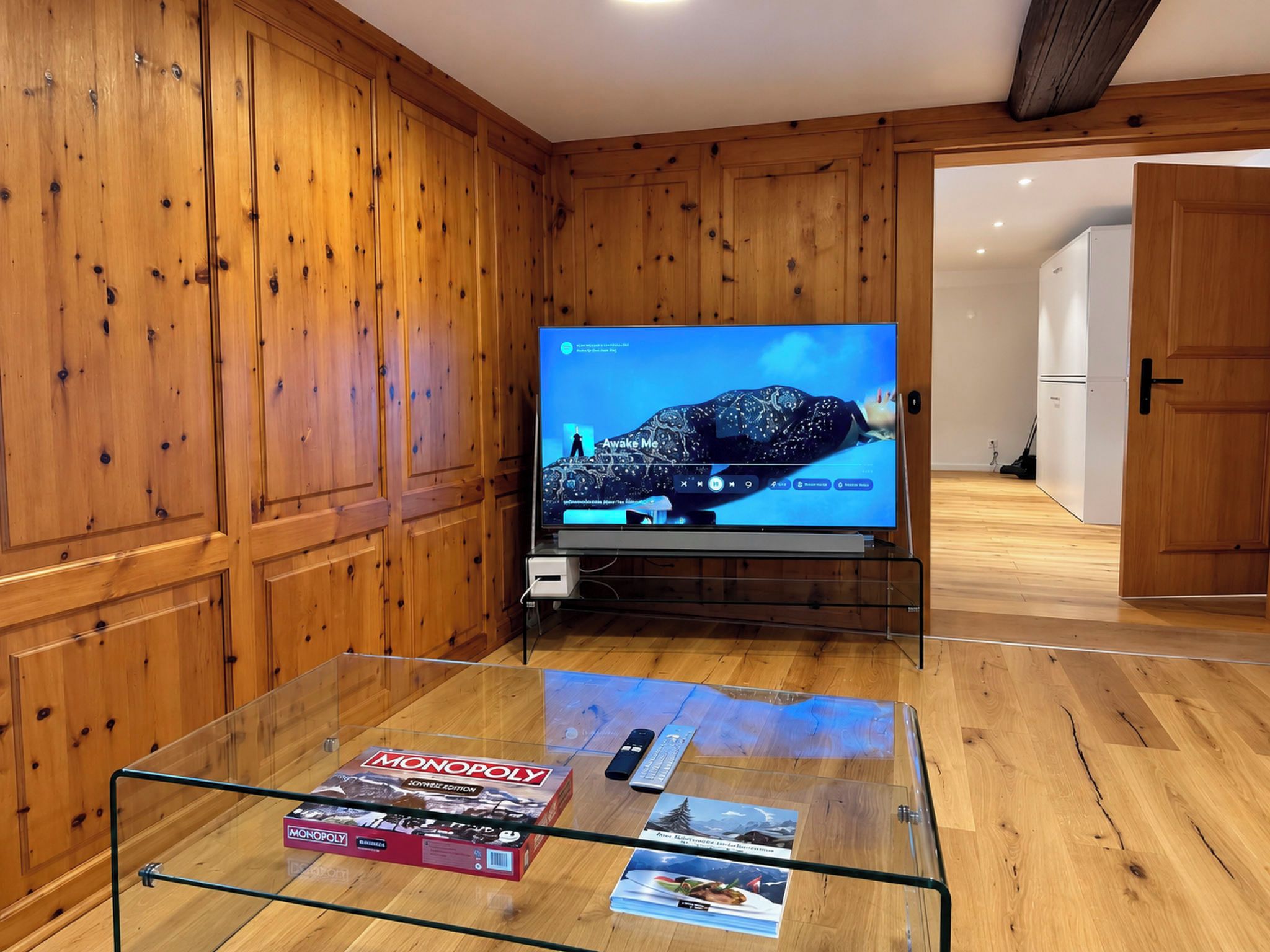 A room with wooden paneling, a television on a stand, and a glass coffee table with a Monopoly game box and magazines.