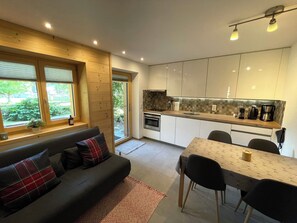 Eine moderne Küche mit weißen Schränken, ein Essbereich mit Tisch und Stühlen sowie eine Sitzecke mit Blick ins Freie.