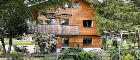 Een houten huis met twee verdiepingen en een balkon, omgeven door een tuin met verschillende planten en een kas.