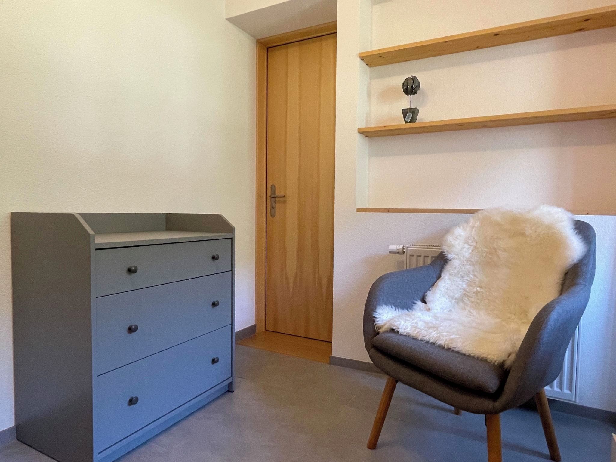 A room with a grey dresser, a wooden door, and a chair with a fluffy white cover.