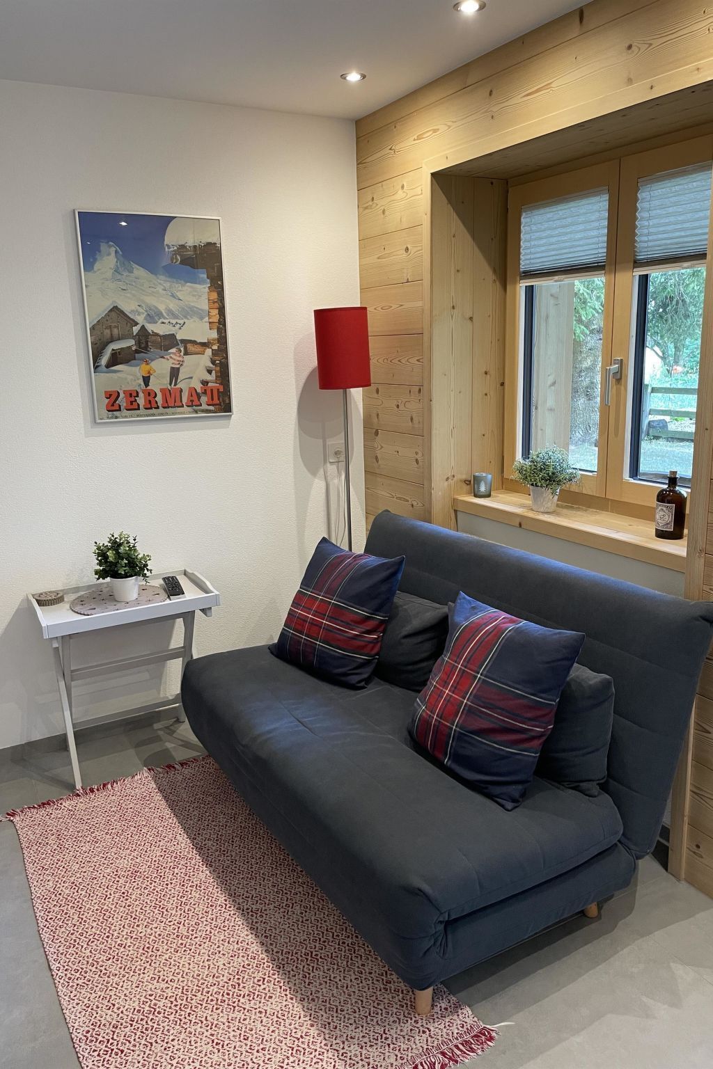 Un salon moderne avec un canapé gris foncé, deux coussins à carreaux, une lampe de sol rouge, une petite table avec une plante et une affiche accrochée au mur.