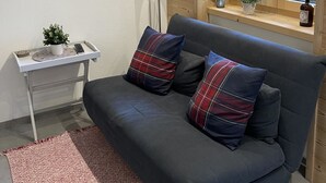 Ett modernt vardagsrum med en mörkgrå soffa, två kuddar i rutigt mönster, en röd golvlampa, ett litet bord med en växt och en affisch på väggen.