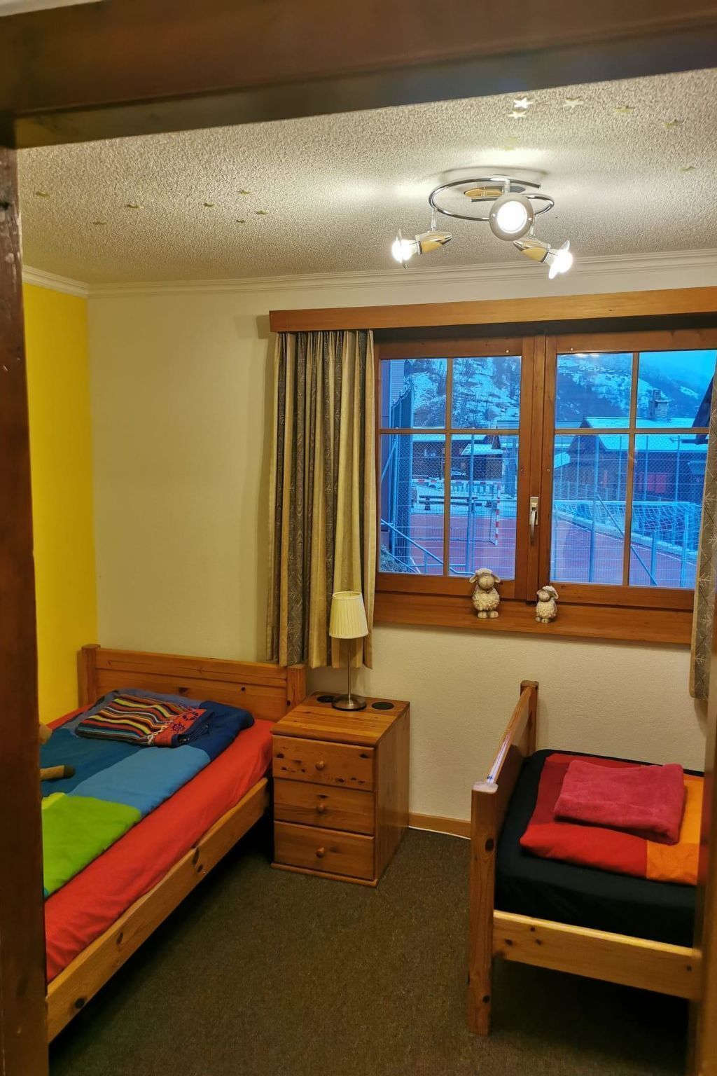 Ein Schlafzimmer mit zwei hölzernen Betten, einem kleinen Holzschrank, einer Lampe und einem Fenster mit Blick auf eine verschneite Landschaft.