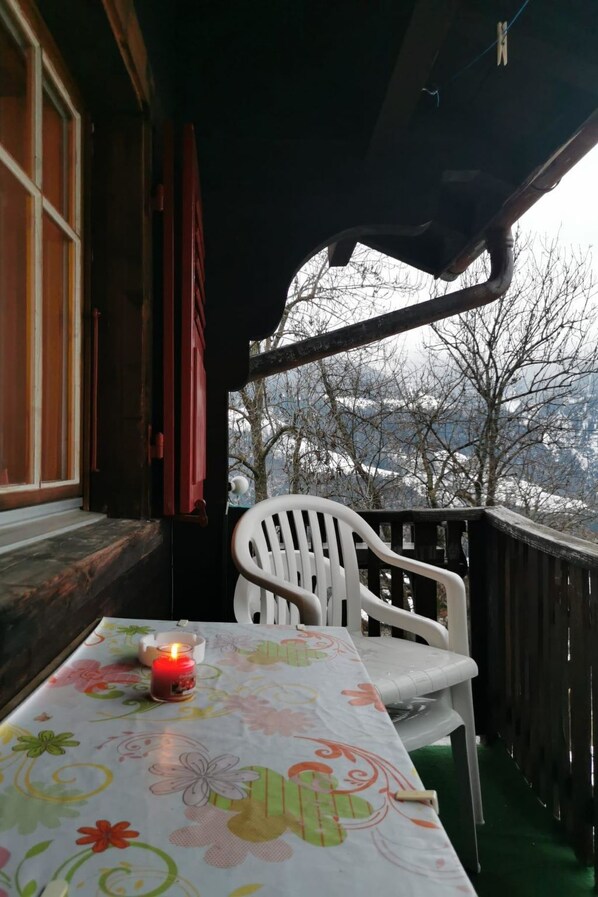 A table with a floral tablecloth and a lit candle, a white plastic chair, and a view of a snowy landscape through a wooden railing.