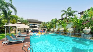 Piscina con sillones de playa, sombrillas y plantas tropicales.