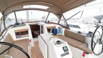 A boat's interior with a steering wheel, control panel, cushioned seating, and a view of the marina.