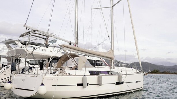 Un yate blanco amarrado en un puerto deportivo con múltiples mástiles y aparejos, rodeado de aguas tranquilas y montañas a lo lejos.