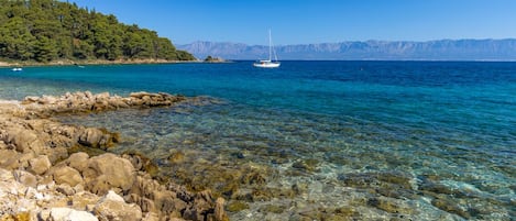 Una costa rocosa con agua cristalina, un velero y una colina boscosa al fondo.
