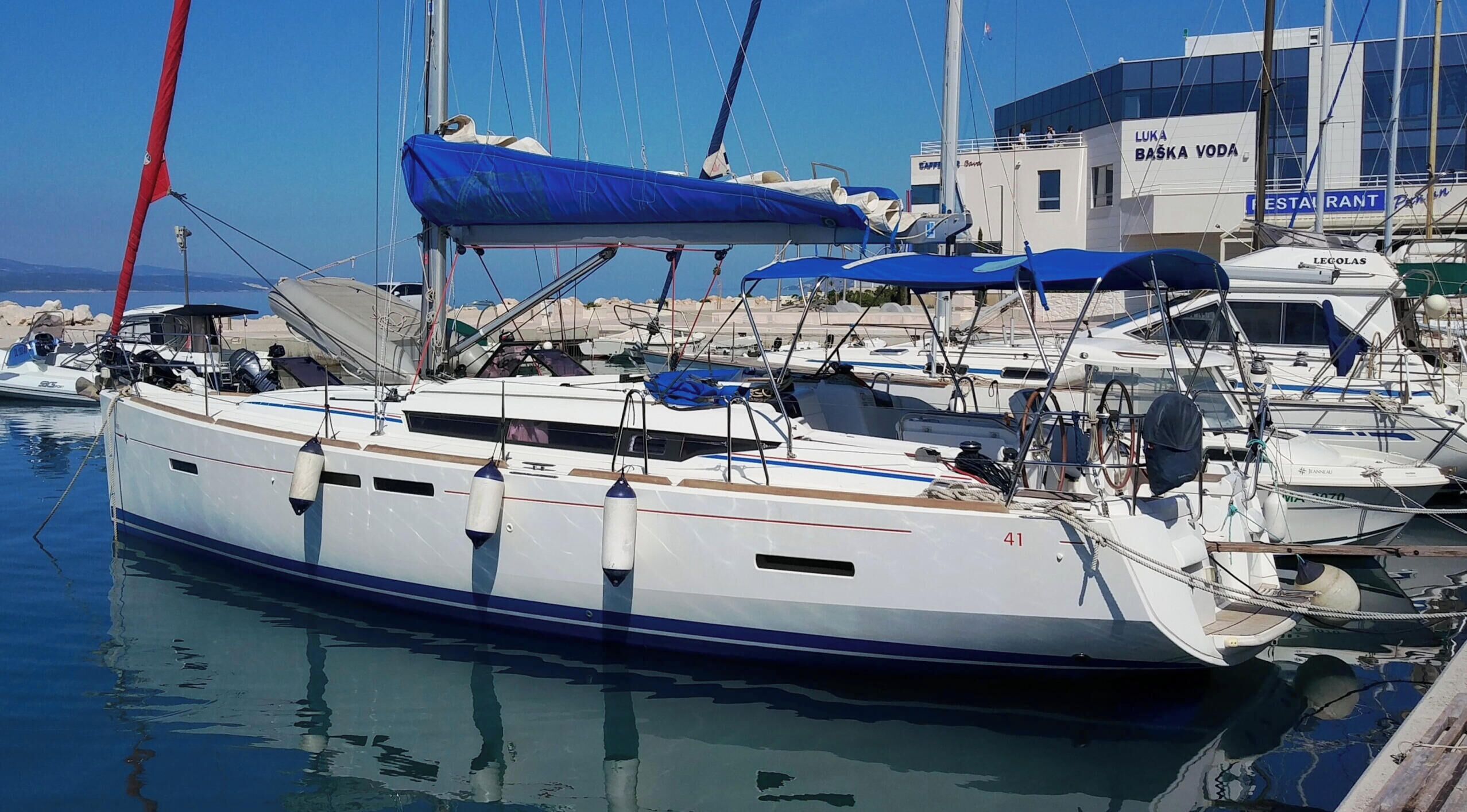Eine Marina mit mehreren dort vertäuten Segelbooten, im Hintergrund ein Gebäude mit der Aufschrift 'LUKA BASKA VODA RESTAURANT' und ein klarer, blauer Himmel.