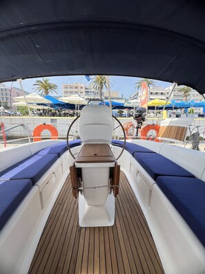 Cubierta de barco con asientos acolchados azules, una mesa central y anillos salvavidas naranjas.