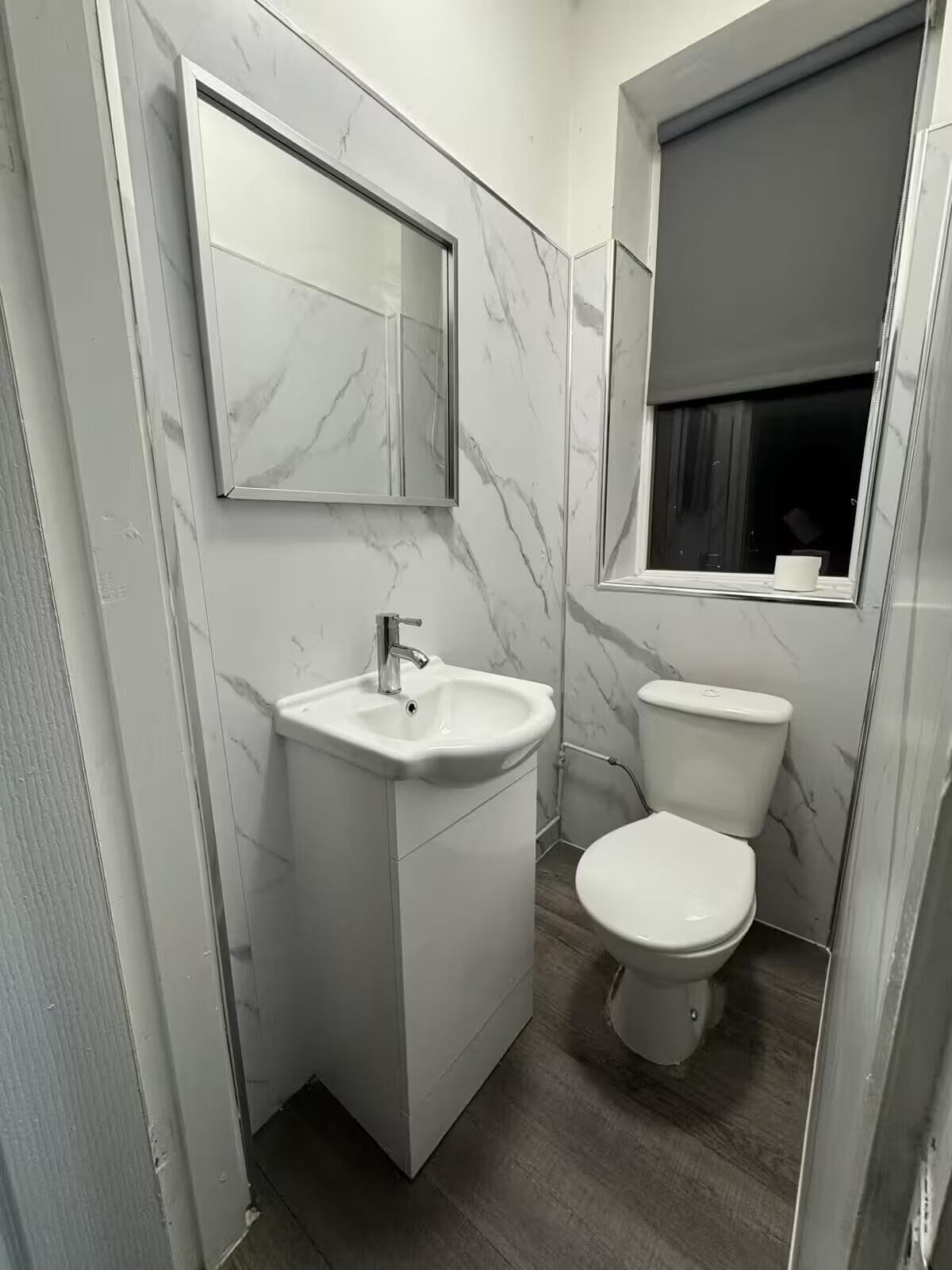 A bathroom with a white sink, mirror, and toilet, marble-patterned walls, and a window with a blind.