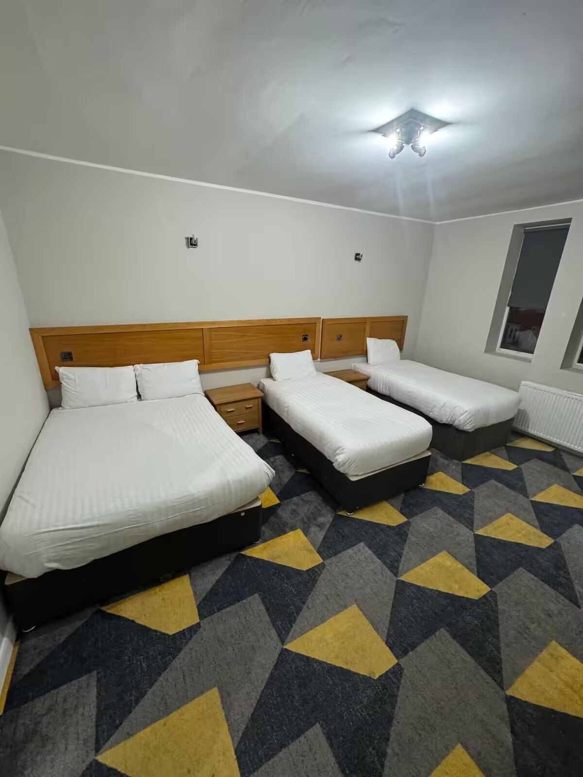 A hotel room with a large bed, two smaller beds, a wooden headboard, a patterned carpet, and a window with blinds.