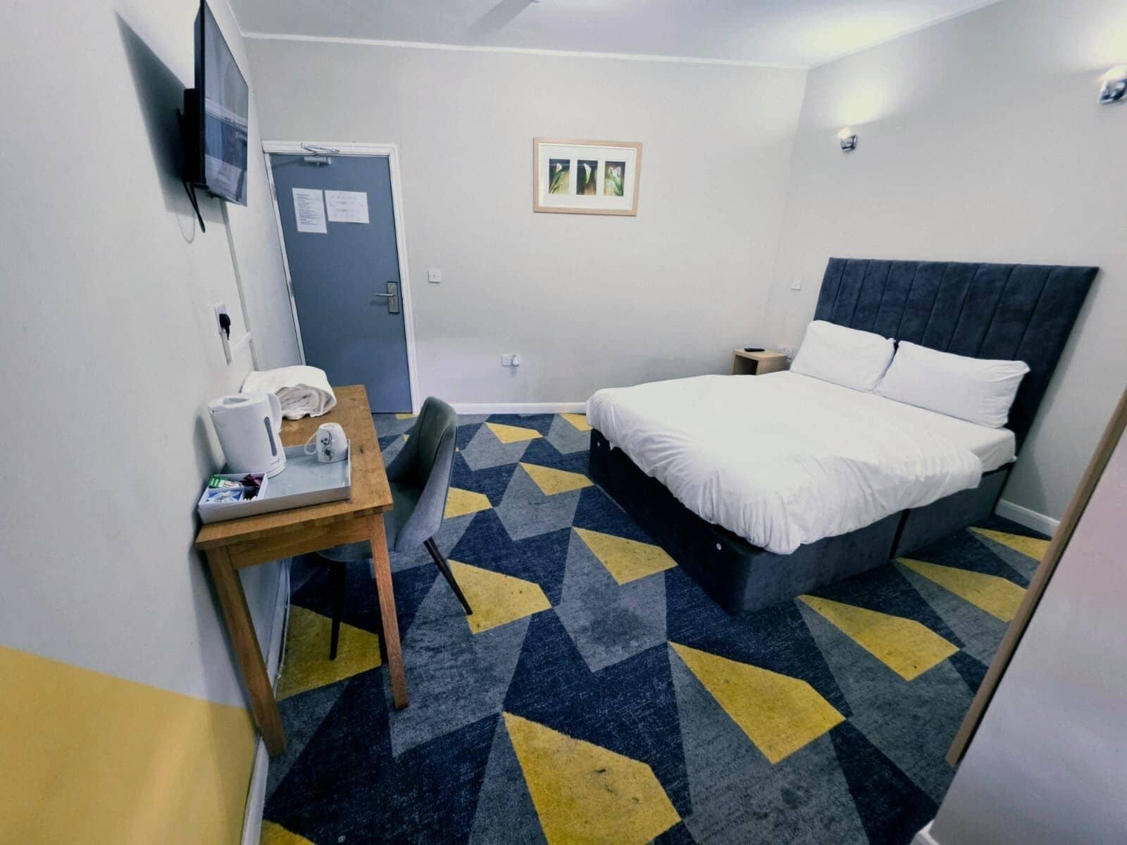 A hotel room with a bed, a desk with a chair, a TV mounted on the wall, and a door with a notice.